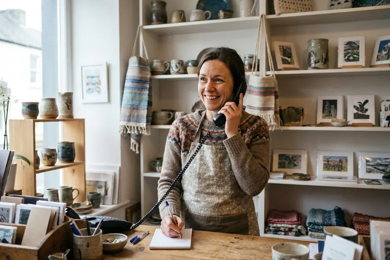 A smiling independent shop owner takes a phone order and writes notes in a craft shop.